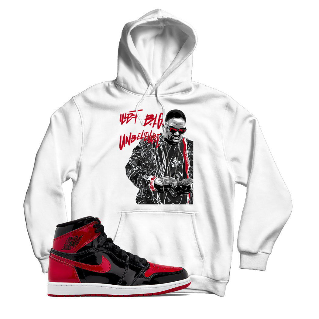 White hoodie with a graphic print of a person wearing sunglasses and a hat, paired with a sneaker matching the Jordan 1 Patent Bred shoe.